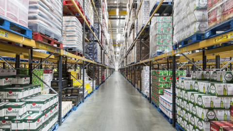 High‑bay warehouse aisle filled with stacked pallets of boxed goods and products organised on multi‑level racking.